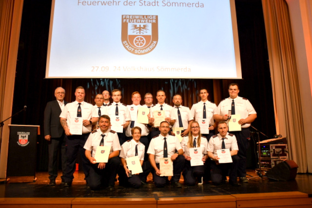 Auf der Bühne im Volkshaus stehen und knien die Ausgezeichneten und Beförderten Feuerwehrmänner und zwei Feuerwehrfrauen in jeweils einer Reihe und halten ihre Urkunden vor der Brust. Auch der Bürgermeister, der Stadtbrandmeister sowie der stellvertretende Kreisbrandinspektorhaben sich eingereiht.