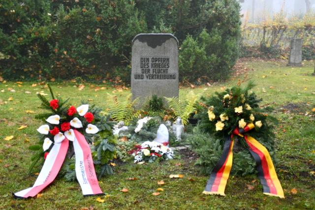 Zwei Kränze mit Blumen und Schleifen liegen links und rechts eines Gedenksteins. Auf diesem steht folgendes: Den Opfern des Krieges, der Flucht und Vertreibung. Direkt vor dem Gedenkstein liegt ein Blumengesteck.