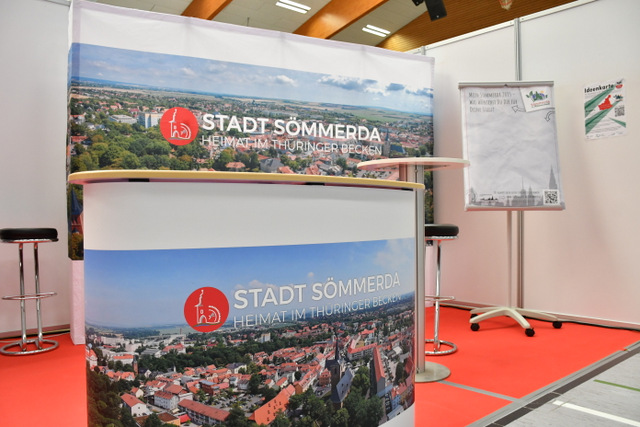 Der Messestand der Stadt Sömmerda mit einem Counter, der an der Frontseite mit einem Luftbild von Sömmerda bespannt ist. Außerdem gibt es dahinter eine große Leinwand mit einem weiteren Luftbild von Sömmerda sowie ein Flipchart, auf dem Besucherinnen und besucher ihre Ideen zum künftigen Sömmerda aufschreiben können.