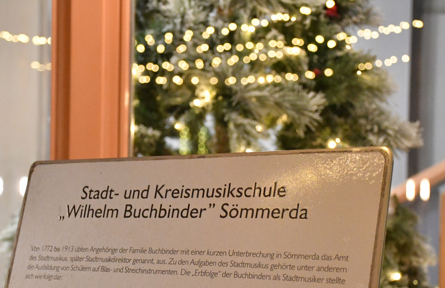 Das Namensschild der Stadt- und Kreismusikschule "Wilhelm Buchbinder" mit einigen kurzen Daten zur Familie Buchbinder.  Angehörige der Familie hatten von 1772 bis 1913 mit einer kurzen Unterbrechung in Sömmerda das Amt des Stadtmusikus, später Stadtmusikdirektor genannt, inne. Hinter dem Schild steht ein geschmückter Weihnachtsbaum mit Kugeln und Lichterkette.