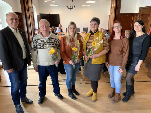 Zwei Männer und vier Frauen stehen in einer Reihe. Die drei gewählten Mitglieder des Vorstandes halten jeweils einen Blumenstrauß in der Hand. Eingerahmt werden sie vom Bürgermeister sowie zwei Vertreterinnen der Abteilung Soziales der Stadtverwaltung. Im Hintergrund sitzen die anderen Mitglieder des Seniorenbeirates an zum Quadrat zusammengeschobenen Tischen und schauen in Richtung der Fotografierten.