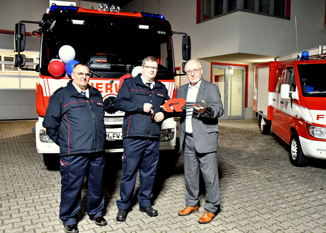 Drei Männer stehen aufgereicht vor dem großen neuen Einsatzfahrzeug. Ein weiteres Feuerwehrfahrzeug steht auf dem Gelände der Feuerwache in Leubingen. Dahinter das erleuchtete Gebäude der Feuerwache. Der Bürgermeister übergibt einen großen symbolischen Schlüssel an den Wehrleiter, dcer neben ihm steht. Dieser hält den kleinen Fahrzeugschlüssel für das HLF. Der dritte Mann vor dem Fahrzeug - er steht neben dem Wehrleiter - ist der stellvertretende Wehrleiter von Leubingen.