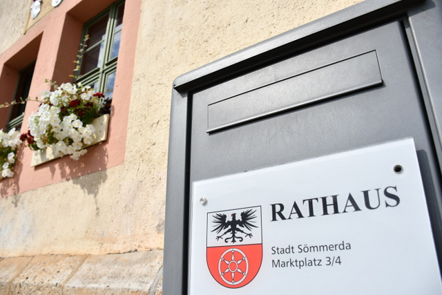 Ein Ausschnitt aus der Vorderansicht des Rathauses Sömmerda mit einem Teil der Fassade und zwei Fenstern, in denen Blumenkästen stehen. Im Vordergrund steht der obere Teil der Briefkasten- und Informationssule vor dem Rathaus. Daran ist ein Schilde mit dem Wappen von Sömmerda und der Aufschrift Rathaus sowie der Adresse angebracht.