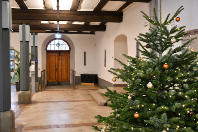 Blick von Innen durchs Rathausfoyer zur geschlossenen Eingangstür. Rechts steht ein mit Kugeln und Lichterkette geschmückter Weihnachtsbaum. Links sind zwei der historischen Pfeiler zu sehen, die die Decke stützen. Sie sind hellgrau, am oberen Ende befinden sich moderne Lampen. Ganz links am Bildrand ist der Teil einer Grünpflanze sichtbar.