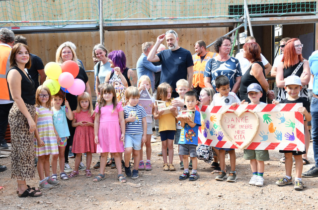 Kindergartenkinder und Erzieherinnen stehen in einer Reihe und halten Luftballons, ein selbst gestaltetes Plakat sowie sowie aus Holzbausteinen selbst gebaute Häuder. Auf dem Plakat steht in einem Herz "Danke für unsere neue Kita". Neben dem Herz sind farbige Handabdrücke der Kinder. Hinter den Kindern stehen lose in einer Gruppe Frauen und Männer. Es sind unter anderem Vertreter der Stadtverwaltung, Bauleute und Planer. Dahinter ist ein Teil des Rohbaus sichtbar.