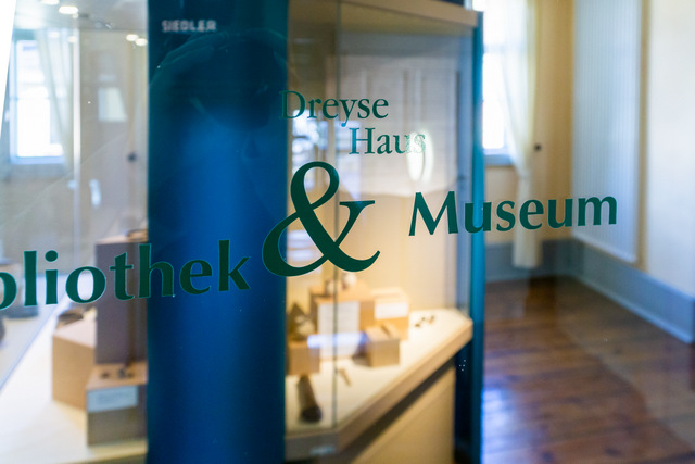 Durch eine Glastür, auf der in der Mitte der Schriftzug "Dreyse Haus Bibliothek & Museum" steht, schaut man in einen Teil des Sonderausstellungsraums des Museums. Kurz hinter der Glastür steht eine blaue Säule, dahinter sind leicht unscharf gemacht Ausstellungsvitrinen mit verschiedenen Objekten zu sehen.