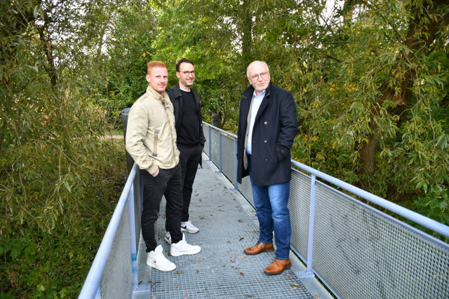 Drei Männer stehen in einer Gruppe auf dem mit einem aus verzinktem Stahl bestehenden langen und schmalen Steg. Der Steg hat ein Geländer ebenfalls aus verzinktem Stahl. Im Hintergrund der Männergruppe gibt es Bäume und Sträucher.