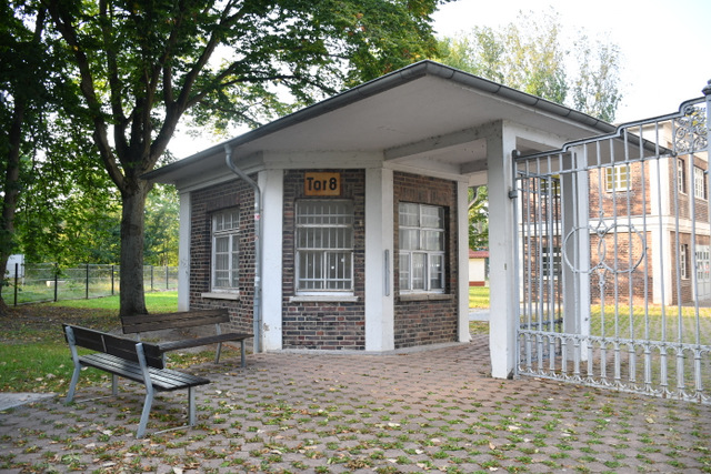 Das sechseckige Torhaus aus braun-roten Ziegelsteinen steht neben einem großen Eingangstor. Die mehrteiligen Fenster sind vo innen mit weißen Gittern versehen. Über einem der Fenster steht auf einem alten Schild Tor 8. Vor dem Torhaus stehen im rechten Winkel zwei Bänke. Zur Seite des Eingangsbereiches des Tores ist das Dach das Torhauses lang gezogen und überdeckt den hier befindlichen Eingangsbereich auf das Gelände von Berufsschule und Kreisvolkshochschule.