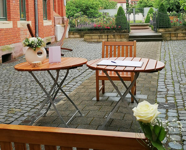 Im schön gestalteten Innenhof des dreiseitigen Dreyse-Hauses stehen eine mit eienr Rose geschmückte Holzbank, zwei runde Holztische - einer davon mit gefüllter Blumenvase. Zwei Bücher liegen auf dem Tisch. Im hintergrund ist hinter einem kunstvollen Zaungitter der Rosengarten mit seinen zahlreichen Rosen und Buchskegeln zu sehen.